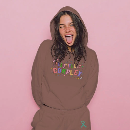 Person wearing a brown hoodie with 'BEAUTIFULLY COMPLEX' text on a pink background